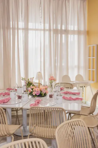 Adult table set with roses and pink napkins beside the floor-to-ceiling curtained windows