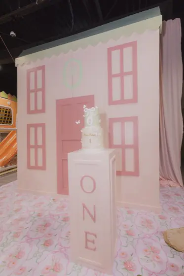Pink playhouse backdrop with the birthday cake on a lettered ONE pedestal