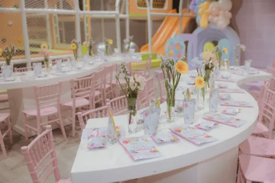 Curved kids' table with pink chiavari chairs, floral bud vases and the indoor playground behind